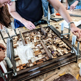Een hout mozaïek in aanbouw van kleine stukjes hout. Met een bruine lijst en plankjes   om iets op te zetten. Gemaakt tijdens een Wall-Art teambuilding.