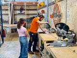 Tijdens de ouder en kind workshop helpt een begeleider met het opmaat zagen van een houten plank.